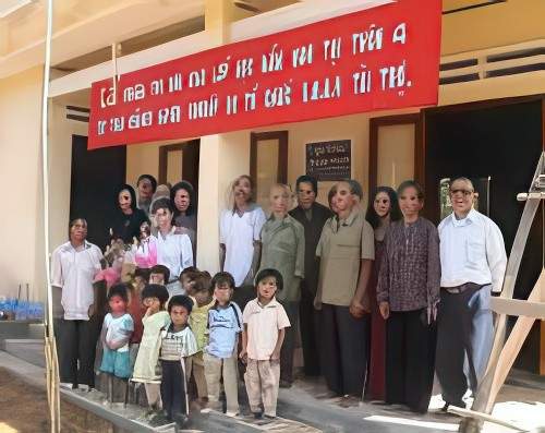 A group of adults and children stand in front of a building adorned with a large red sign. The children are positioned at the front, while the adults stand behind them.