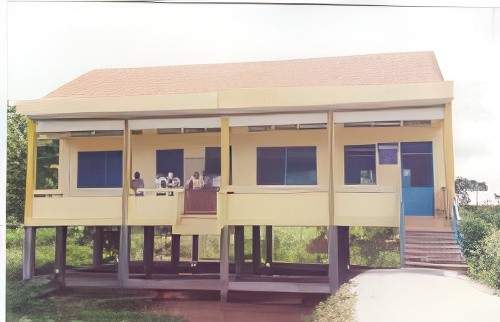 A yellow elevated building with a red roof, featuring three large windows and a blue door with stairs leading up to it. There are four people visible on the porch. The background includes greenery.