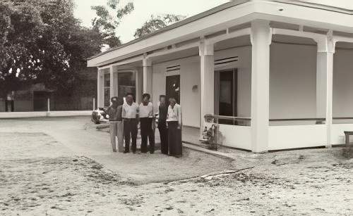 A sepia-toned photograph of a single-story building with a gable roof and multiple columns at the front. The building is surrounded by open, barren land.