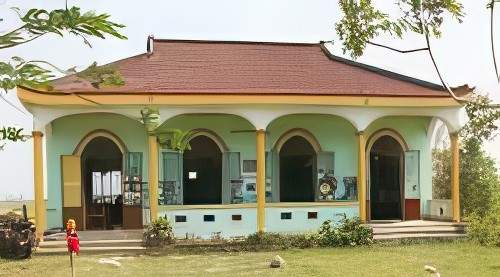 A small green building with a red roof and three archways. A child in a red outfit stands near the entrance, and two motorcycles are parked to the left. Trees surround the building.