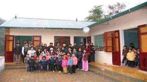 A teacher stands behind five children in a sparsely furnished classroom with earthen walls and a wooden ceiling.