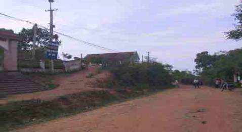 A dirt road with sparse vegetation on either side, power lines overhead, and a few buildings in the background. A sign on a pole is visible on the left side of the image.