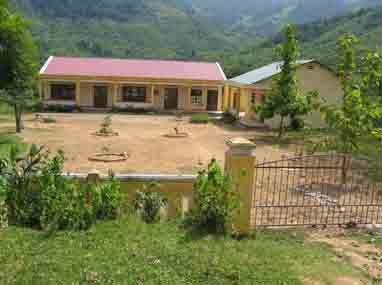 Front view of a one-story building with a red roof, located in a rural area surrounded by greenery and mountains in the background. A fenced area is visible in the foreground.