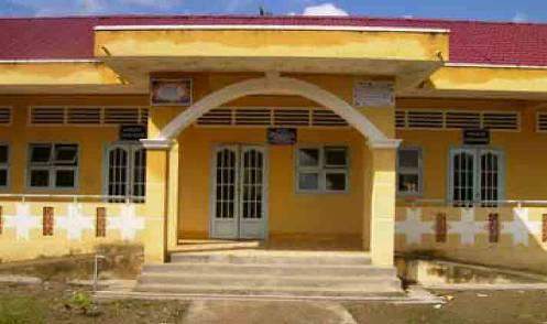 A yellow building with a red roof and arched entrance, featuring closed double doors and a few windows, showing signs of wear. There are steps leading up to the entrance.