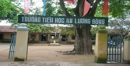 The image shows the entrance gate of Truong Tieu Hoc An Luong Dong, a school with a green sign and some trees in the background. The gateposts are yellow and white, and several students are visible inside.