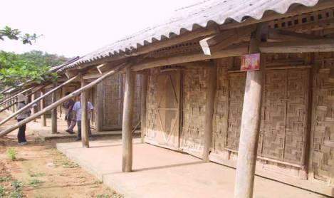 A row of thatched buildings stands beside a dirt path. Several wooden supports prop up the roof. A tree and some greenery are visible in the background on the left side.