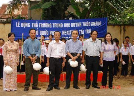 A group of six adults stands in front of children holding white hats, with a blue banner in the background.