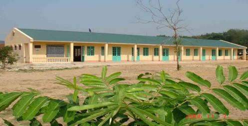 A single-story building with green doors and windows stands in an open area with sparse vegetation. A small tree is in the foreground.