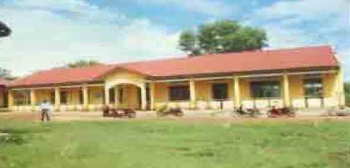 A single-story yellow building with a red roof surrounded by green grass and trees under a partly cloudy sky.