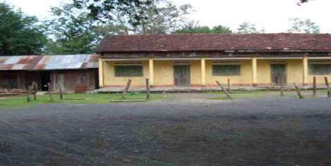 A single-story building with a yellow facade and red roof, surrounded by trees, with an open dirt space and wooden pole barriers in front.