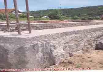 A concrete foundation slab with metal rods protruding upwards, set in a construction site with a hill and greenery in the background.