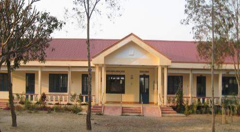 A one-story building with a red roof, beige walls, and a central entrance, surrounded by trees and shrubs.