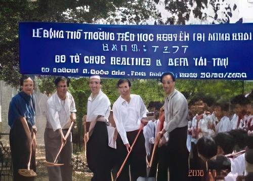 A group of people stand under a blue banner during a groundbreaking ceremony, holding shovels and smiling. Children are seated to the right. The event appears to be taking place outdoors.