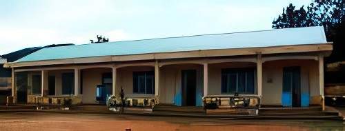 A single-story building with a metal roof and several blue doors, situated on dirt ground. The structure has a columned front and is surrounded by a rural landscape with trees.