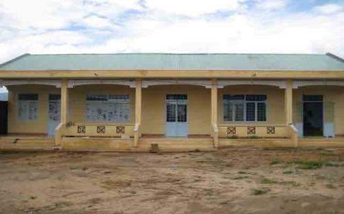 A small, single-story building with three doors and windows. It has a beige exterior and a metal roof, set against a cloudy sky backdrop. The ground in front is uneven and barren.