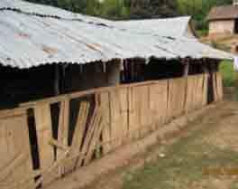 A long, rustic wooden structure with a corrugated metal roof, set in a rural area. The building shows signs of wear and damage. Trees and another building are visible in the background.