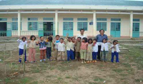 A group of children and two adults stand posing in front of a building with large windows, lined up in two rows. The ground is bare and the building has a teal and white facade.