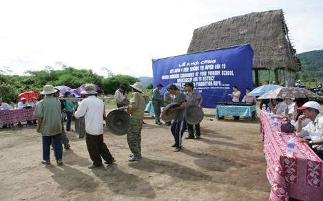 A group of people engage in a traditional dance outdoors, with spectators sitting under umbrellas. A large blue sign with text is displayed in the background near a structure with a thatched roof.