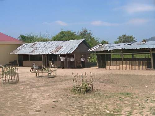 A rural outdoor area with two simple structures, one with a metal roof and another with a thatched roof. Vegetation and a partly cloudy sky are visible in the bac