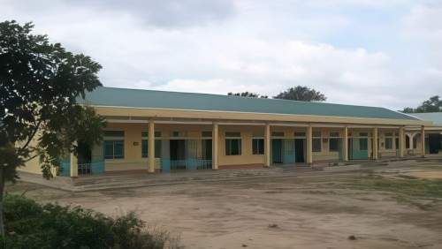 A single-story yellow building with a green roof and several doors and windows. The structure is surrounded by a dirt ground and some greenery, with a cloudy sky in the background.