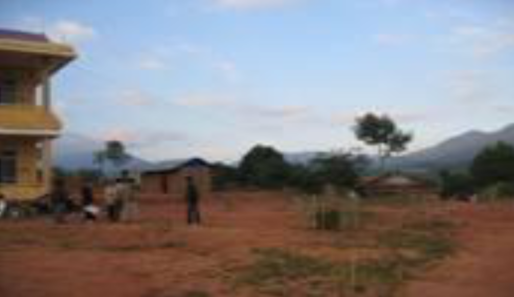 A landscape with red soil, scattered trees, and a few buildings, including a yellow multi-story structure. People are walking and gathering near the buildings under a blue sky with clouds.