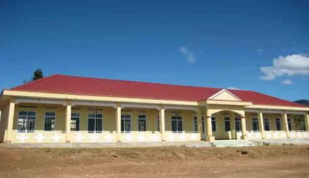 A long, single-story building with a red roof and yellow walls, featuring multiple windows and columns in the front. The building is set against a clear blue sky with a few scattered clouds.