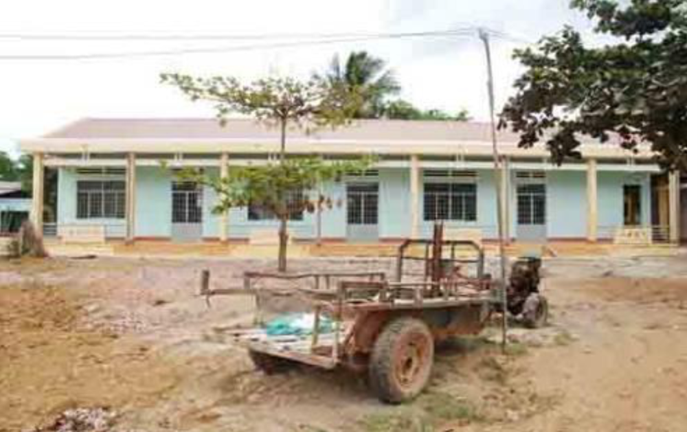 A single-story light blue building with a brown roof stands behind an old, rusty suspension frame of a vehicle in a yard with patchy grass.