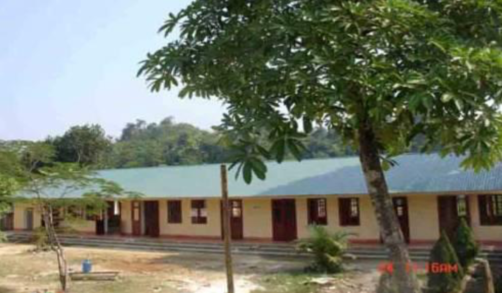 A single-story building with a green roof and several doorways is partially obscured by trees. The ground is light brown and appears sandy or dry. The building is surrounded by greenery.