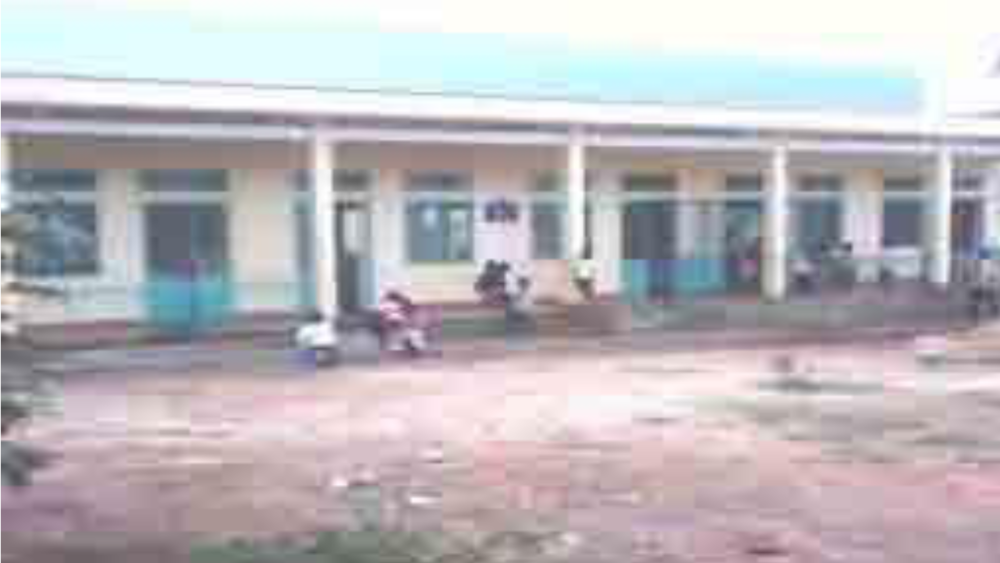 A row of single-story buildings with a few people sitting and standing outside. The ground in front is bare and unpaved.