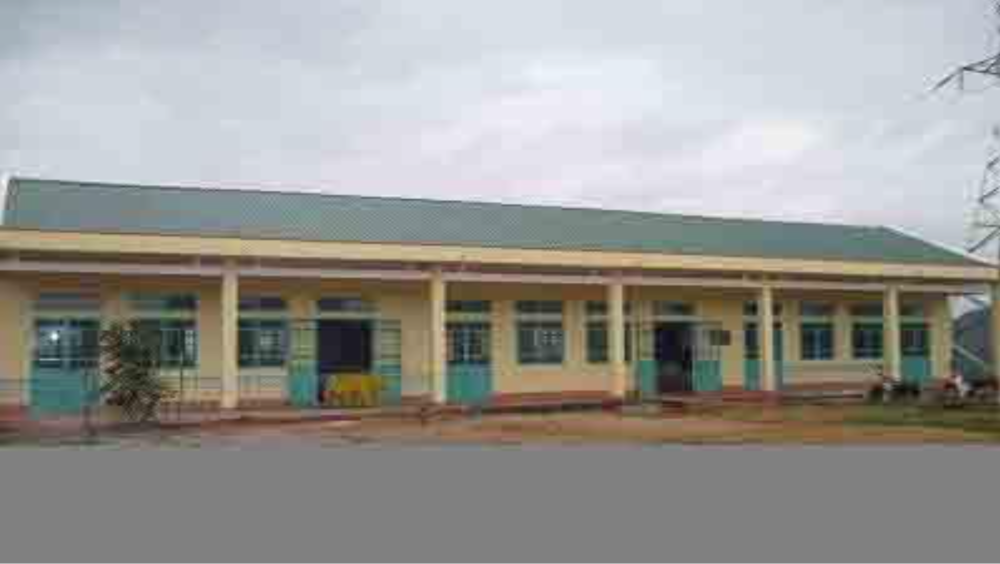 A single-story building with a green roof and cream-colored walls, featuring multiple blue-framed windows and doors. The structure is surrounded by a paved area and sparse vegetation.