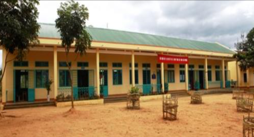 A single-story yellow building with a green roof and multiple blue doors and windows, surrounded by a few trees and a dirt courtyard. A red sign is displayed above the central entrance.