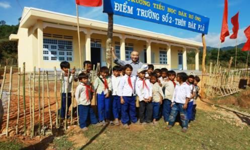 A group of children in school uniforms stand with an adult in front of a building with a sign written in Vietnamese. Red flags are visible in the background.