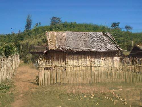 A small, thatched-roof house stands in a rural area surrounded by a bamboo fence, with a dirt path leading to the entrance. A hill with greenery is visible in the background under a clear blue sky.