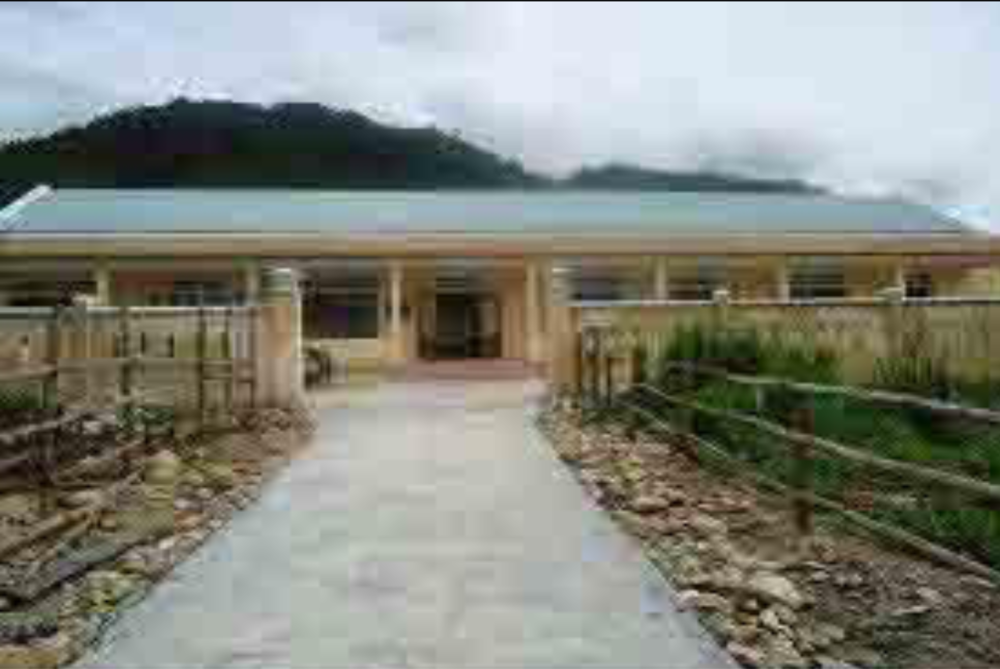 A one-story building with a light blue roof is set against a hilly landscape. A concrete path leads to the entrance, which is flanked by wooden fences on either side.