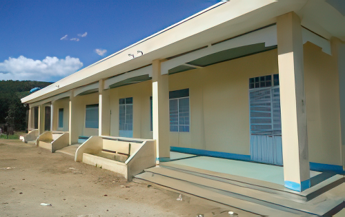 A single-story building with yellow walls and a long porch supported by pillars. The structure has multiple doors and windows, and the ground outside is bare soil.