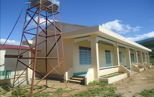 A beige building with a metal tower beside it, situated under a clear blue sky. The area around the building appears dry.