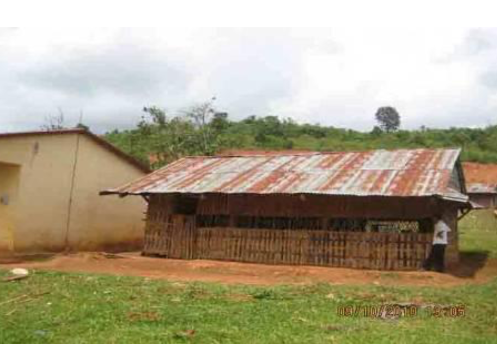 A small building with a rusted tin roof and an open structure made of wooden planks in a grassy area next to a cream-colored structure. Trees and hills are visible in the background.