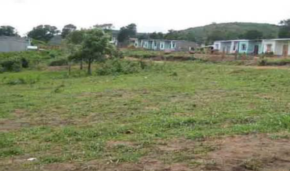 A grassy field with scattered houses and trees in the background under a cloudy sky.