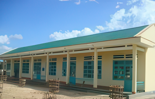 A small, single-story wooden house elevated on stilts under a clear blue sky with some clouds. The surrounding area is grassy with some trees in the background.