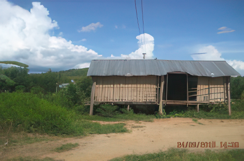 A small, single-story wooden house elevated on stilts under a clear blue sky with some clouds. The surrounding area is grassy with some trees in the background.