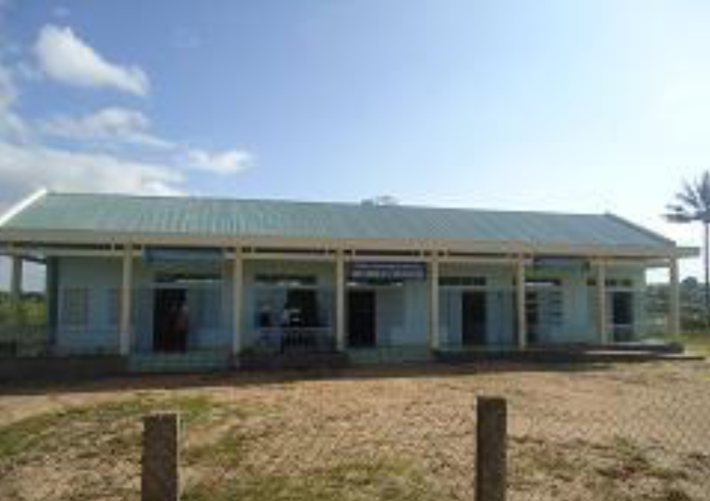 Single-story building with light blue walls, numerous windows, and a green roof, situated behind a wire fence, on a sunny day.