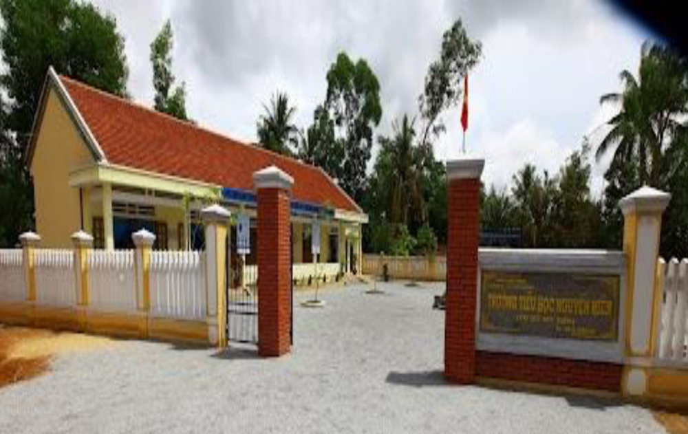 Two single-story, yellow buildings with red-tiled roofs sit on a dirt lot surrounded by trees and a blue sky. One building is smaller and simpler, while the larger one has a covered walkway.