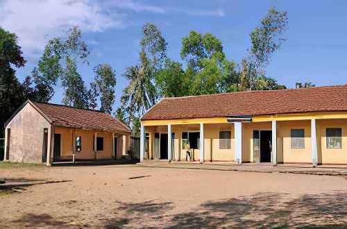 Two single-story, yellow buildings with red-tiled roofs sit on a dirt lot surrounded by trees and a blue sky. One building is smaller and simpler, while the larger one has a covered walkway.