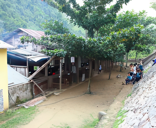 A small group of people sits along a sloped hillside near a row of modest buildings and trees in a rural area.