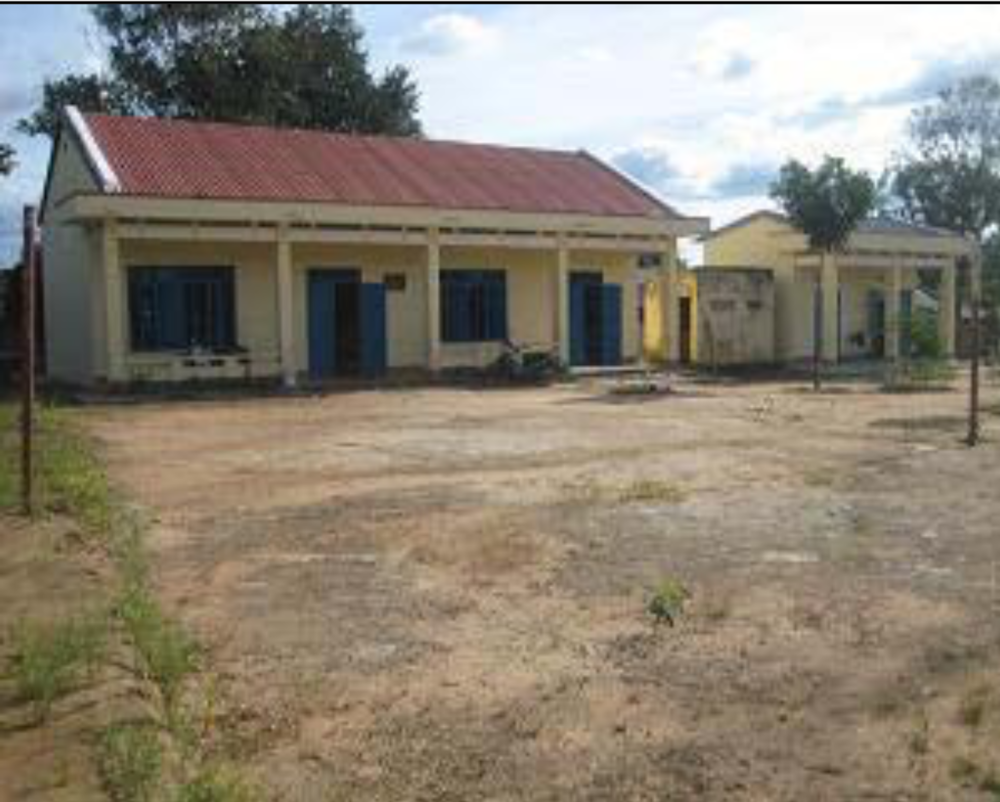 A school building with a sign above the entrance reading "TRUONG TIEU HOC LIEN SON DIEM TRUONG SO 2 THON DAKRIJOP." The area around the school appears rural with cloudy skies in the background.