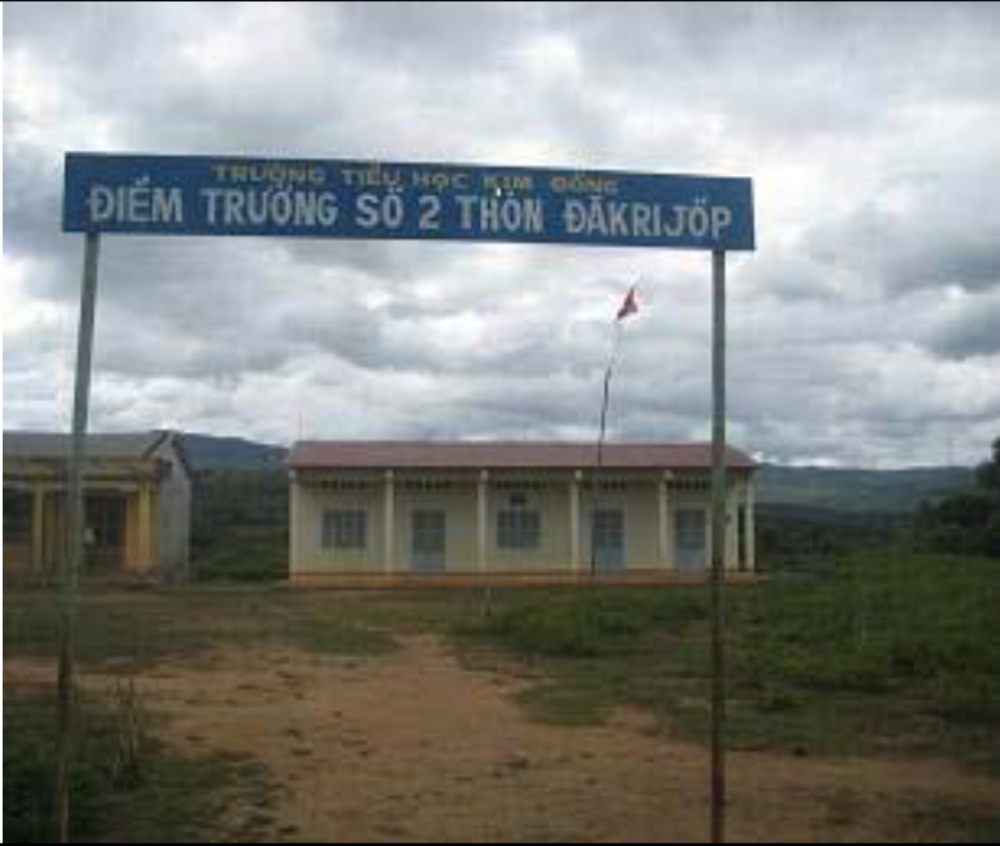 A school building with a sign above the entrance reading "TRUONG TIEU HOC LIEN SON DIEM TRUONG SO 2 THON DAKRIJOP." The area around the school appears rural with cloudy skies in the background.