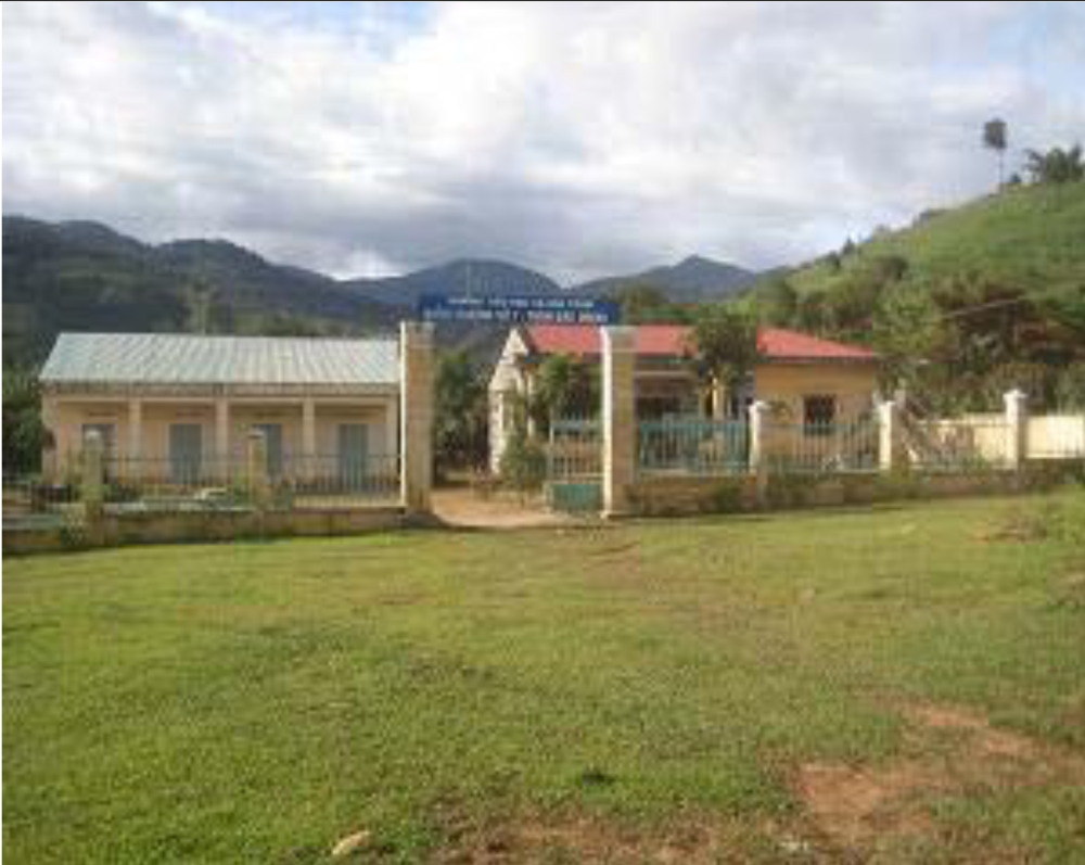 A small building complex with a sign in front, located in a grassy area with mountainous terrain in the background under a partly cloudy sky.