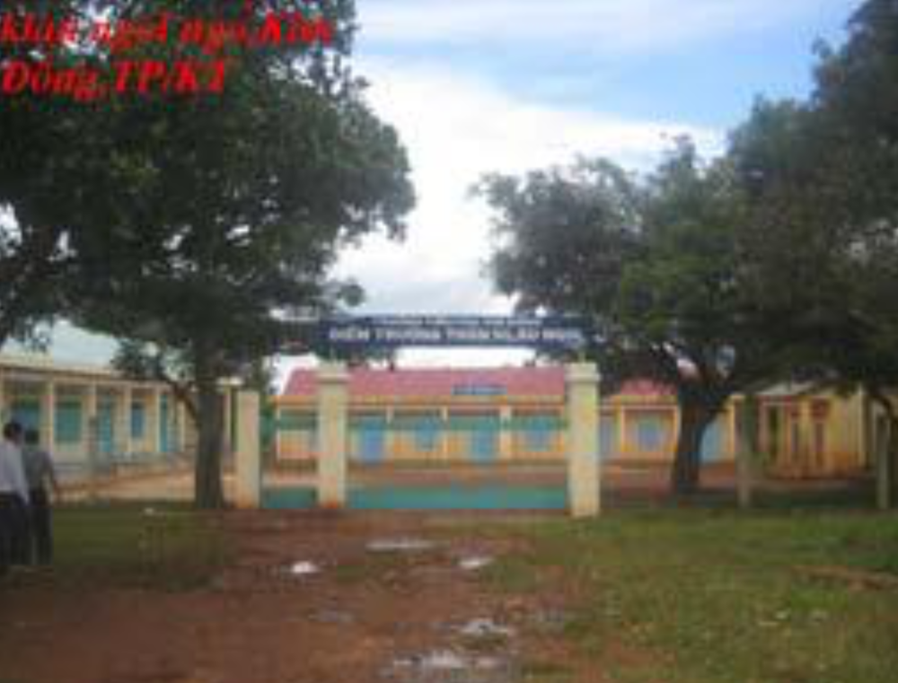 A single-story building with a red roof and light-colored walls, featuring several blue doors and windows. It is set on a brown dirt ground with trees visible on one side.