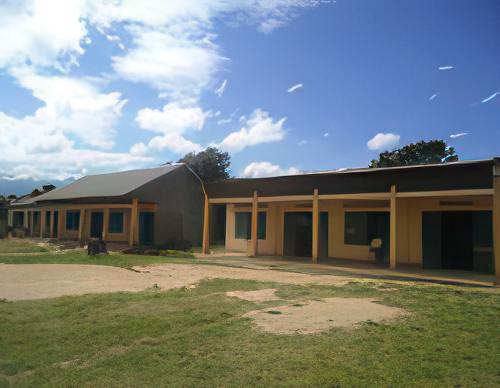 A single-story building with beige walls and blue doors, surrounded by grass and a clear blue sky with scattered clouds.