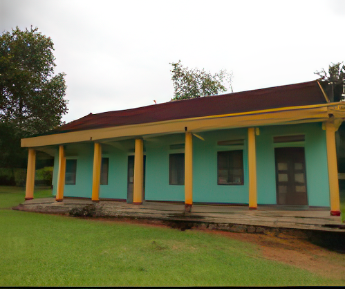 A single-story building with a teal facade, yellow columns, and a red roof sits on a grassy area with trees in the background.